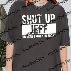 May include: Dark gray t-shirt with the text "SHUT UP JEFF" in large white letters. Below the name is the phrase "NO MORE FROM YOU TODAY". The shirt is a casual style.