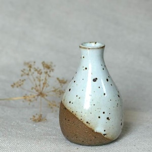 May include: A small ceramic bud vase with a narrow neck and a rounded base. The upper portion features a glossy white glaze with dark brown speckles, while the lower section is finished in a textured, unglazed earthy brown clay finish.