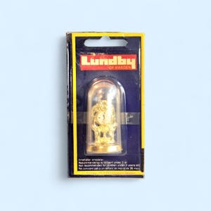 May include: A gold-colored miniature clock inside a clear dome, packaged on a blue and yellow card. The card features the brand name "Lundby of Sweden" and text in multiple languages, including "Not recommended for children under 3 years old."