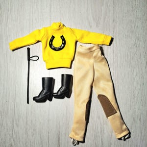 Op de afbeelding: Een miniatuur paardensportoutfit met een gele coltrui met een zwart hoefijzerontwerp, beige rijbroek met bruine knielappen, zwarte rijlaarzen en een zwarte rijzweep. De outfit is ontworpen voor een pop.