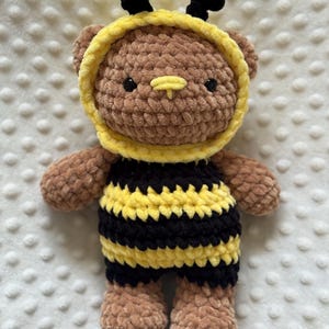 May include: A brown crocheted bear plush toy wearing a yellow and black striped bee costume. The bear has black safety eyes, a small yellow stitched nose, and a yellow hood with two black antennae on top. The body features horizontal stripes.