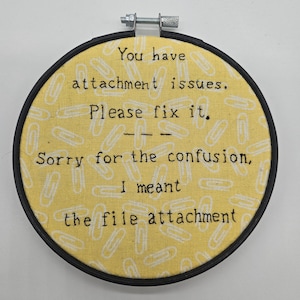 Può includere: Un telaio da ricamo rotondo con uno sfondo in tessuto giallo con disegni di graffette bianche. Il testo recita: "Hai problemi di attaccamento. Per favore, risolvilo. Scusa la confusione, intendevo l'allegato."