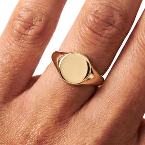 May include: A gold signet ring with a smooth, circular face is worn on a finger. The ring is a warm, shiny gold colour. The background is plain white.