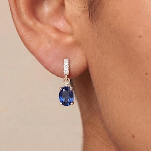 May include: A gold-coloured earring featuring a blue oval gemstone and a small bar of white gemstones. The earring is displayed on a person's ear, with the focus on the jewellery.
