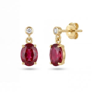 Könnte beinhalten: Ein Paar goldene Ohrstecker, jeweils mit einem leuchtenden, oval geschliffenen Rubin. Ein kleiner, runder Diamant akzentuiert die Oberseite jedes Ohrrings. Die Ohrringe sind in einem klassischen, eleganten Design gehalten.