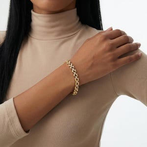 Op de afbeelding: Een goudkleurige armband met een herhalend bloemmotief. De armband wordt om de pols van een persoon gedragen. De armband heeft een dubbelrijig ontwerp met kleine, heldere stenen in het bloemmotief. De persoon draagt een beige coltrui.