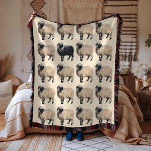 May include: A woven throw blanket featuring a pattern of black and white sheep. The blanket has a fringed border and is being held up by a person.