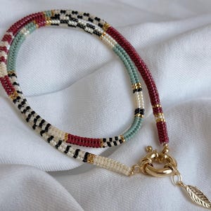 May include: A beaded necklace with a gold clasp and feather charm. The necklace features a pattern of red, white, black, and gold beads. The necklace is displayed on a white, textured surface.