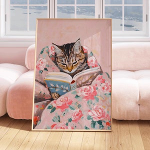 Puede incluir: Una ilustración enmarcada de un gato leyendo un libro mientras está acurrucado en una manta con estampado floral. El gato es marrón y atigrado con ojos verdes. El fondo es de color rosa suave.