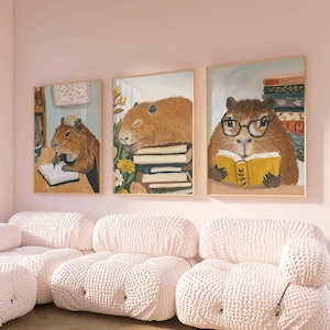 May include: Three framed illustrations of capybaras, each with a different activity. One capybara is writing in a notebook, another is sleeping on a stack of books, and the third is reading a book while wearing glasses. The illustrations are hung on a pink wall above a white modular sofa.
