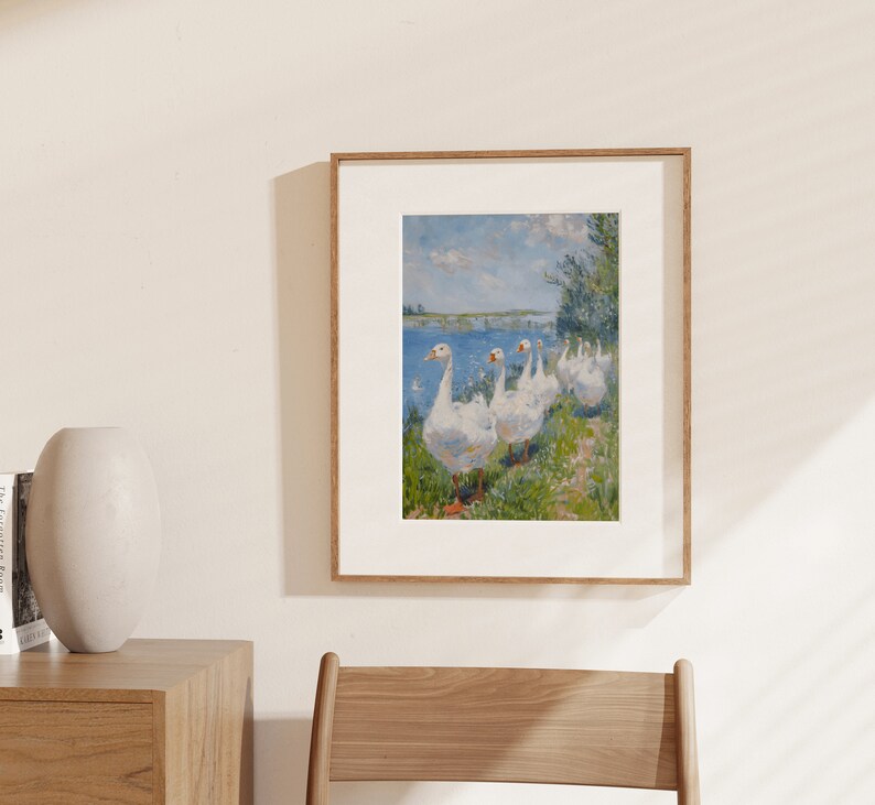May include: A framed painting of a group of white geese walking along a grassy path by a body of water. The geese are in a line, with the one at the front looking directly at the viewer. The painting is done in a realistic style, with a focus on the details of the geese's feathers and the surrounding landscape.