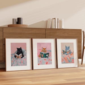 Op de afbeelding: Drie ingelijste illustraties van katten die in bed boeken lezen. Elke kat heeft een andere kleur: zwart, oranje en grijs. De illustraties zijn op een roze achtergrond met bloemenpatronen.