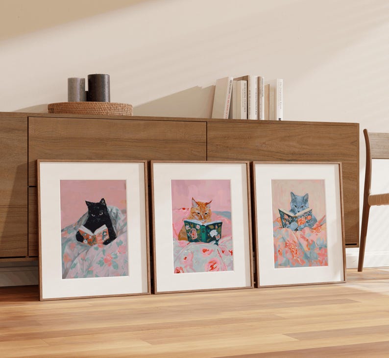 Op de afbeelding: Drie ingelijste illustraties van katten die in bed boeken lezen. Elke kat heeft een andere kleur: zwart, oranje en grijs. De illustraties zijn op een roze achtergrond met bloemenpatronen.