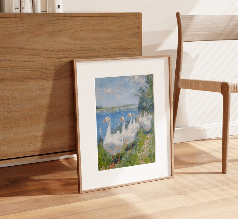 May include: A framed print of a painting depicting a group of white geese walking in a line on a grassy bank by a body of water. The geese are facing the right side of the image. The background is a blue sky with white clouds and a green field.