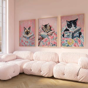 Op de afbeelding: Drie ingelijste illustraties van katten die boeken lezen terwijl ze in dekens met bloemenpatronen liggen. De illustraties hangen aan een lichtroze muur boven een witte modulaire bank.