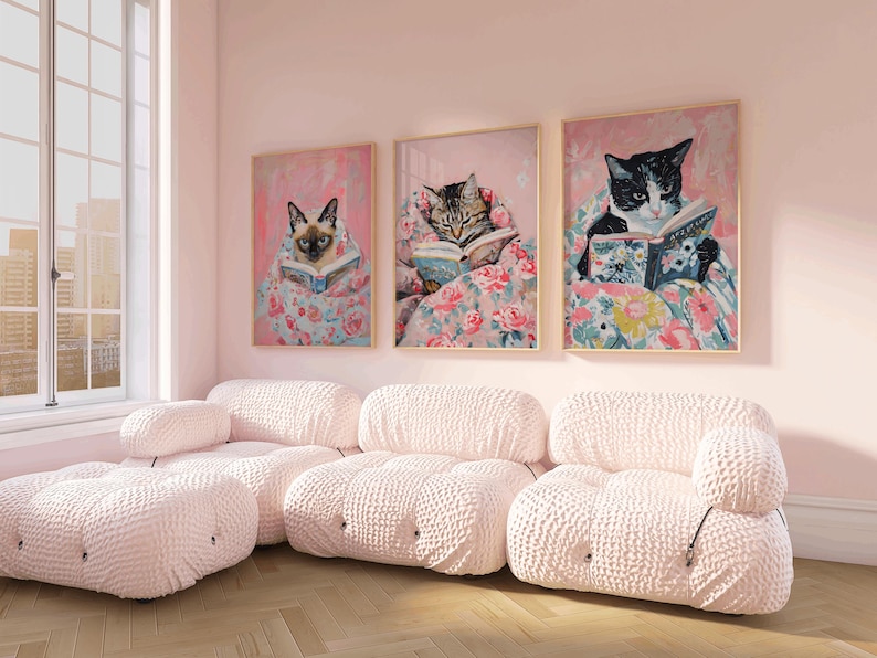 Op de afbeelding: Drie ingelijste illustraties van katten die boeken lezen terwijl ze in dekens met bloemenpatronen liggen. De illustraties hangen aan een lichtroze muur boven een witte modulaire bank.