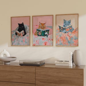 Op de afbeelding: Drie ingelijste illustraties van katten die boeken lezen. Elke kat is in een andere kleur, zwart, oranje en blauw, en elke ligt in een bed met bloemenprint beddengoed. De illustraties zijn tegen een witte muur geplaatst.