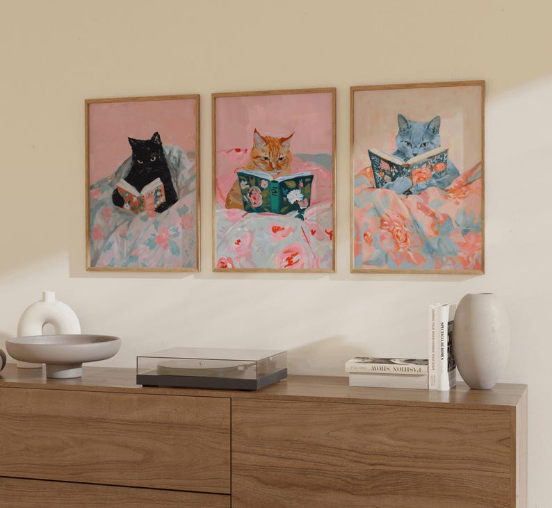 Op de afbeelding: Drie ingelijste illustraties van katten die boeken lezen. Elke kat is in een andere kleur, zwart, oranje en blauw, en elke ligt in een bed met bloemenprint beddengoed. De illustraties zijn tegen een witte muur geplaatst.
