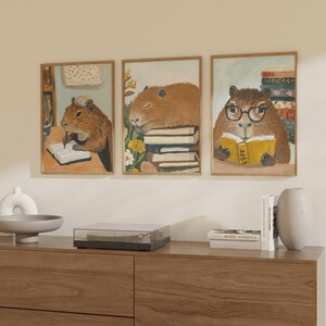 May include: Three framed illustrations of capybaras reading books. The capybaras are brown and have large, round ears. The books are colourful and have different titles. The frames are wooden and have a light brown finish.