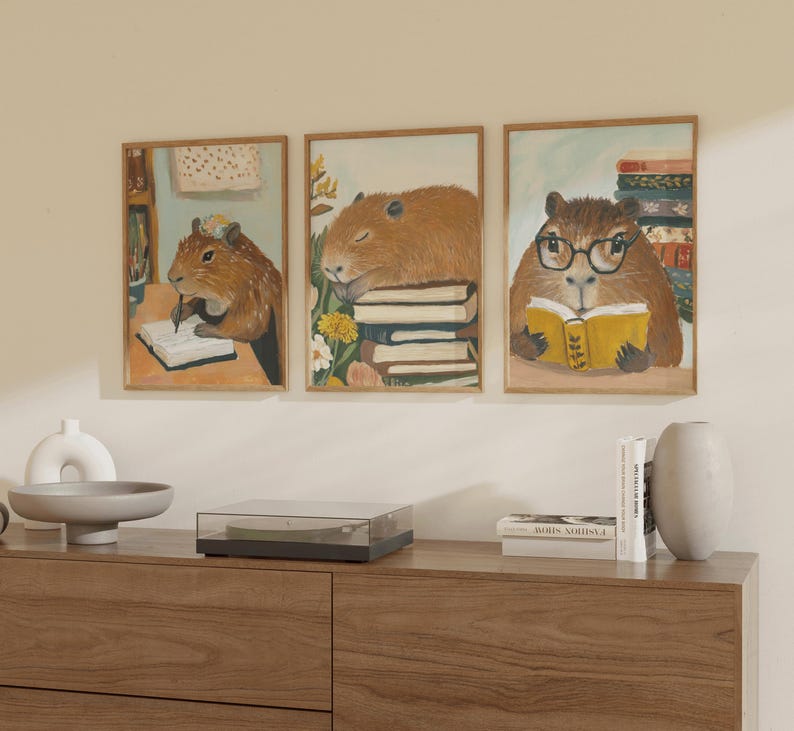 May include: Three framed illustrations of capybaras reading books. The capybaras are brown and have large, round ears. The books are colourful and have different titles. The frames are wooden and have a light brown finish.