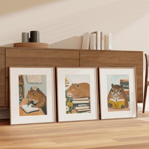 May include: Three framed illustrations of capybaras with books. The capybara on the left is writing in a notebook, the middle capybara is sleeping on a stack of books, and the capybara on the right is reading a book.