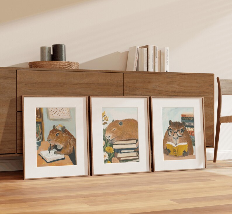 May include: Three framed illustrations of capybaras with books. The capybara on the left is writing in a notebook, the middle capybara is sleeping on a stack of books, and the capybara on the right is reading a book.