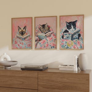 Op de afbeelding: Drie ingelijste prints van katten die in bed boeken lezen. Elke print toont een ander kattenras en een andere boekomslag. De katten liggen allemaal op een roze en bloemig beddengoed.