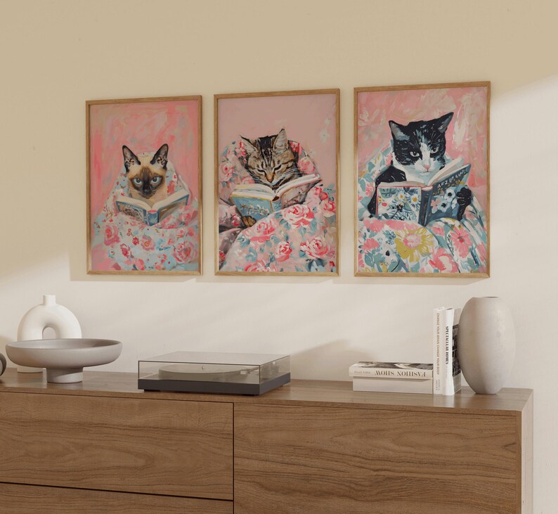 Op de afbeelding: Drie ingelijste prints van katten die in bed boeken lezen. Elke print toont een ander kattenras en een andere boekomslag. De katten liggen allemaal op een roze en bloemig beddengoed.