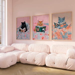 Op de afbeelding: Drie ingelijste schilderijen van katten die in bed boeken lezen. De schilderijen hangen in een roze kamer met een witte modulaire bank.