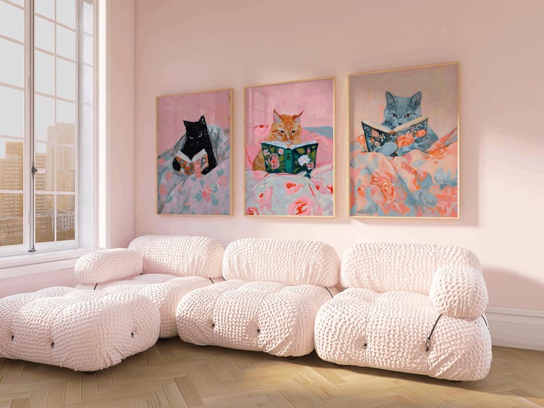 Op de afbeelding: Drie ingelijste schilderijen van katten die in bed boeken lezen. De schilderijen hangen in een roze kamer met een witte modulaire bank.