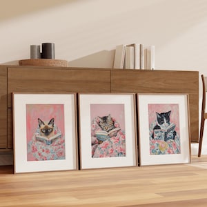 Op de afbeelding: Drie ingelijste prints van katten die boeken lezen. Elke kat ligt in een roze bloemenpatroon deken. De kat links is een Siamese kat, de kat in het midden is een tabby kat en de kat rechts is een zwart-witte kat.