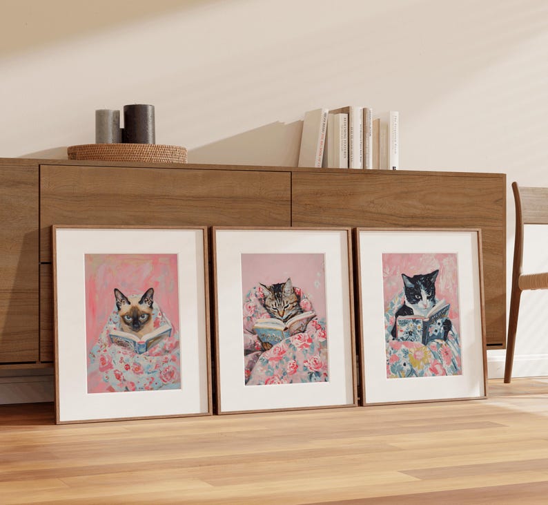 Op de afbeelding: Drie ingelijste prints van katten die boeken lezen. Elke kat ligt in een roze bloemenpatroon deken. De kat links is een Siamese kat, de kat in het midden is een tabby kat en de kat rechts is een zwart-witte kat.