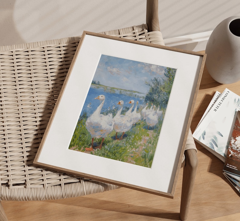 May include: A framed print of a painting depicting a group of white geese walking along a riverbank. The geese are in a line, with their heads down, and their feathers are ruffled. The background is a blue sky with white clouds, and the river is a light blue color. The painting is in a simple wooden frame.