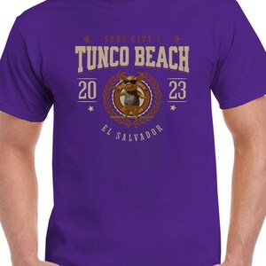 May include: Purple t-shirt with a graphic of a pig wearing sunglasses and a surfboard. The graphic says "Surf City | Tunco Beach 20 23 El Salvador".