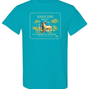 May include: A turquoise blue t-shirt with a graphic print of a pig standing on a beach with palm trees. The graphic has the text "PLAYA EL TUNCO SURF CITY! LA LIBERTAD, EL SALVADOR QUALITY SURF TIME SINCE 2002"