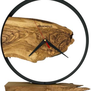 Op de afbeelding: Een wandklok bestaande uit een lichtbruine natuurlijke houten schijf met zichtbare nerven en knoesten, gevat in een dunne, zwarte, cirkelvormige metalen rand. De wijzerplaat toont de tekst PRISEWALD, zwarte wijzers en een rode secondewijzer.