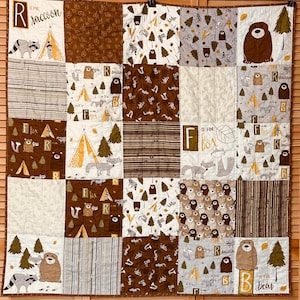 May include: A patchwork quilt featuring woodland animal designs. The quilt has squares with raccoons, bears, foxes, and teepees. The letters "R", "B", and "F" are also featured. The color palette includes brown, white, and tan.