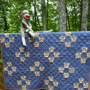 May include: A blue and white quilt with a checkered border. The quilt is made up of squares with a pattern of smaller squares. A sock monkey is sitting on top of the quilt.