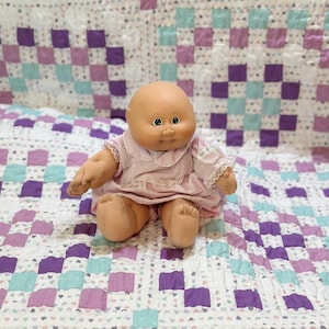 May include: A Cabbage Patch Kid doll wearing a pink dress with lace trim, sitting on a purple and blue patchwork quilt.