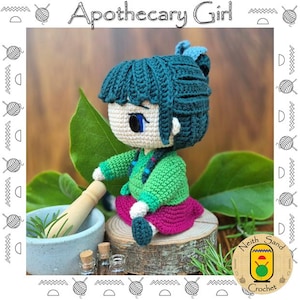 Puede incluir: Muñeca de ganchillo hecha a mano que representa a una chica boticaria. La muñeca tiene cabello verde azulado, una chaqueta verde y una falda rosa. Está sentada junto a un mortero y una maja, con el texto "Apothecary Girl" arriba. La muñeca está sobre una superficie de madera.