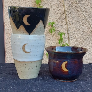 Pode incluir: Um copo alto de cerâmica com uma secção superior preta com uma cordilheira e uma lua crescente, passando para uma base branca salpicada. Ao lado, uma chávena de cerâmica mais curta e de bordo largo em azul escuro com um detalhe de lua.