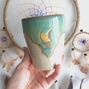 May include: A ceramic mug featuring a two-tone glaze with a glossy teal upper section and a matte beige lower section. The surface is decorated with carved, scalloped mountain-like patterns and a small, yellow crescent moon shape near the rim.