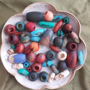 Può includere: Una collezione di perline in ceramica assortite di varie forme, dimensioni e colori, tra cui tonalità di blu, rosso, arancione e marrone. Molte perline presentano motivi botanici o di montagna dipinti a mano in una ciotola di ceramica smerlata.