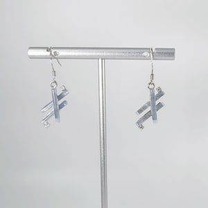 May include: A pair of silver dangle earrings with a geometric design made of blue glass. The earrings are hanging from a silver stand.