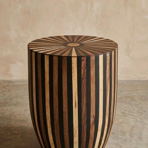 Puede incluir: Una mesa auxiliar cilíndrica con un patrón de rayas verticales que alterna listones de madera natural clara y madera marrón oscura. La parte superior circular muestra un diseño de rayos de sol que irradia desde un círculo central.