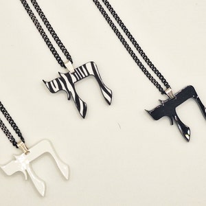 Puede incluir: Tres collares con colgantes Chai. Un colgante es blanco, otro es de estampado de cebra en blanco y negro, y otro es negro liso. Cada colgante cuelga de una cadena negra. Los colgantes son de material brillante.