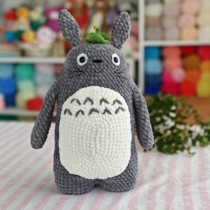 May include: A handmade gray plush toy resembling a character with large eyes, pointed ears, and a white belly patch. It has a small green leaf on its head and is made of a soft, textured material. The toy is standing on a pink and white striped surface.