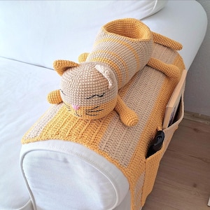 Puede incluir: Organizador de reposabrazos de sofá con forma de gato tejido a ganchillo, en beige y amarillo. El organizador tiene una cabeza de gato, un cuerpo cilíndrico y bolsillos para guardar cosas. La cara del gato tiene los ojos cerrados, una nariz rosa y bigotes negros.