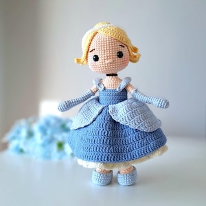 Puede incluir: Muñeca de ganchillo hecha a mano de una princesa con un vestido azul. La muñeca tiene cabello rubio, una diadema blanca y zapatos azules. El vestido es de un azul vibrante con una falda azul más clara. La muñeca tiene un collar negro y piel clara.