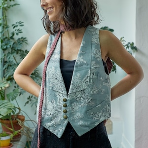 Könnte beinhalten: Eine Frau trägt eine blaue Weste mit einem weißen und grauen Blumenmuster. Die Weste hat einen Schalkragen und vier goldene Knöpfe. Sie trägt auch einen rot-weißen Schal um den Hals.